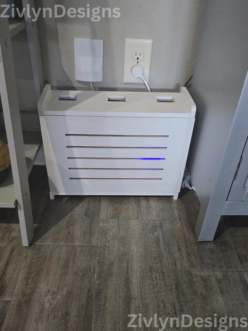 Puede incluir: Una caja de madera blanca con listones horizontales y una luz azul, dise&ntilde;ada para ocultar aparatos electr&oacute;nicos. La caja est&aacute; colocada sobre un suelo con aspecto de madera, con un enchufe de pared y un dispositivo blanco en el fondo.