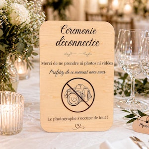 Peut inclure: Panneau en bois clair aux coins arrondis, avec texte en français et symbole de caméra barrée. Le message invite les invités à profiter du moment sans photos ni vidéos, le photographe s'occupant de tout.