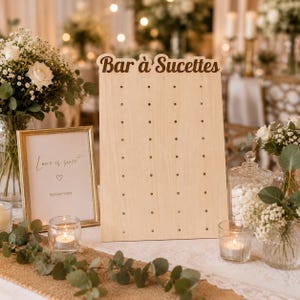 Peut inclure: Un panneau de présentation en bois clair avec le texte Bar à Sucettes en haut et une grille de petits trous pour maintenir des objets. À côté se trouve un cadre avec le texte Love is sweet et Servez-vous, accompagné d'une petite icône de cœur.