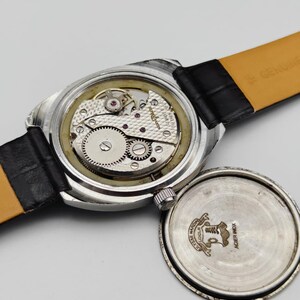 May include: An open-faced vintage wristwatch with a black leather band. The watch mechanism is visible, showcasing intricate gears and jewels. The watch case is silver-toned, and the back cover is detached, revealing the watch's inner workings.