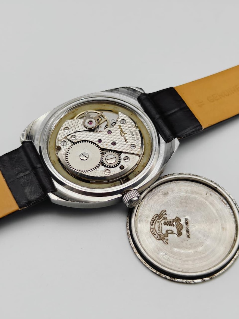 May include: An open-faced vintage wristwatch with a black leather band. The watch mechanism is visible, showcasing intricate gears and jewels. The watch case is silver-toned, and the back cover is detached, revealing the watch's inner workings.