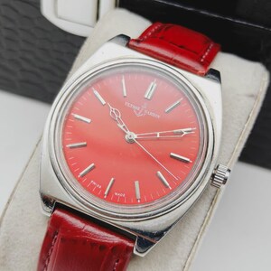 May include: A vintage Ulysse Nardin wristwatch with a vibrant red dial and silver-tone case. The watch features a red leather strap and is marked "Swiss Made." The watch is displayed on a light-colored surface.
