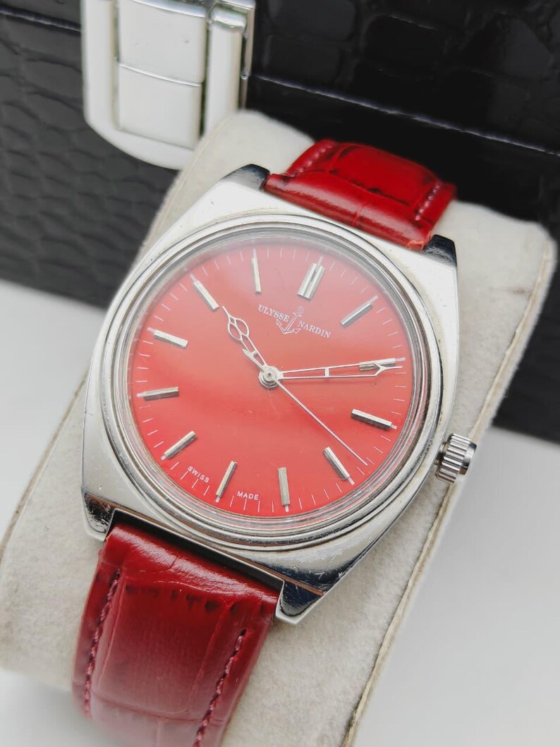May include: A vintage Ulysse Nardin wristwatch with a vibrant red dial and silver-tone case. The watch features a red leather strap and is marked "Swiss Made." The watch is displayed on a light-colored surface.