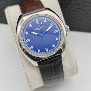 May include: A stainless steel wristwatch with a deep blue face and a brown leather strap. The watch has white hour markers and hands, and the brand name "ULYSSE NARDIN" is visible on the face. The watch is displayed on a white cushion.