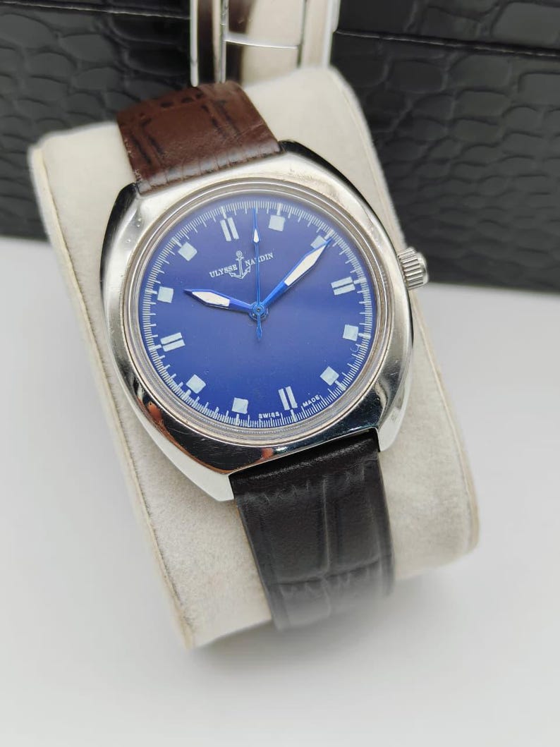 May include: A stainless steel wristwatch with a deep blue face and a brown leather strap. The watch has white hour markers and hands, and the brand name "ULYSSE NARDIN" is visible on the face. The watch is displayed on a white cushion.