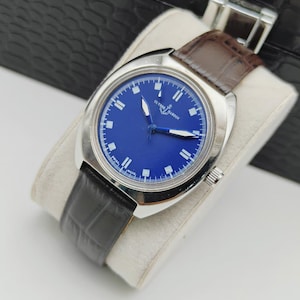 May include: A vintage Ulysse Nardin watch with a striking blue dial and silver-tone case. The watch features white hour markers and hands, complemented by a brown leather strap. The watch is displayed on a white cushion.