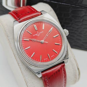 May include: A vintage Ulysse Nardin wristwatch with a vibrant red dial and silver-tone case. The watch features silver hour markers and hands, and a red leather strap. The watch is displayed on a white cushion.