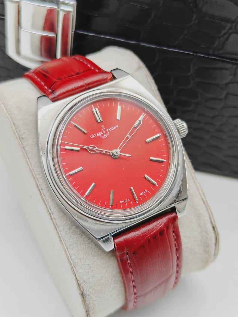May include: A vintage Ulysse Nardin wristwatch with a vibrant red dial and silver-tone case. The watch features silver hour markers and hands, and a red leather strap. The watch is displayed on a white cushion.