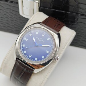 May include: A vintage Ulysse Nardin watch with a blue dial and silver case. The watch features a brown leather band and is displayed on a white cushion. The watch is set against a black, textured background.
