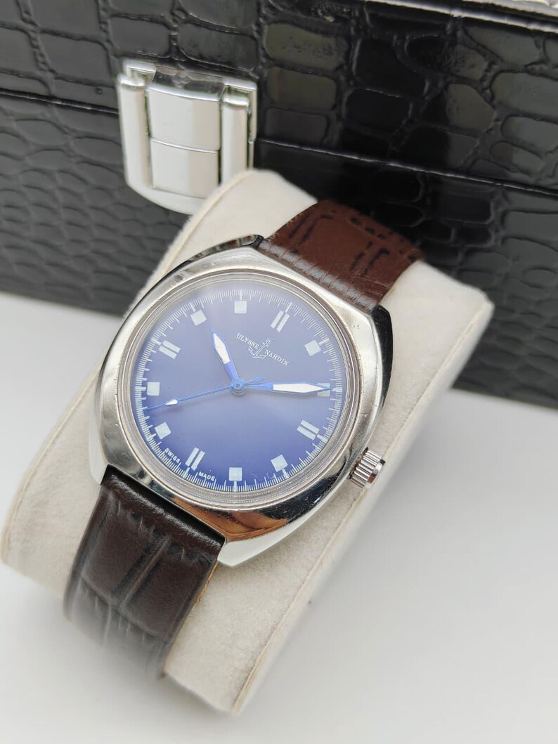 May include: A vintage Ulysse Nardin watch with a blue dial and silver case. The watch features a brown leather band and is displayed on a white cushion. The watch is set against a black, textured background.
