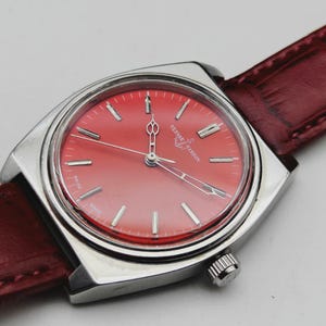 May include: A vintage Ulysse Nardin wristwatch with a vibrant red dial and silver-tone case. The watch features a red leather band and a classic design. The dial displays silver hour markers and hands. The watch is a stylish accessory.