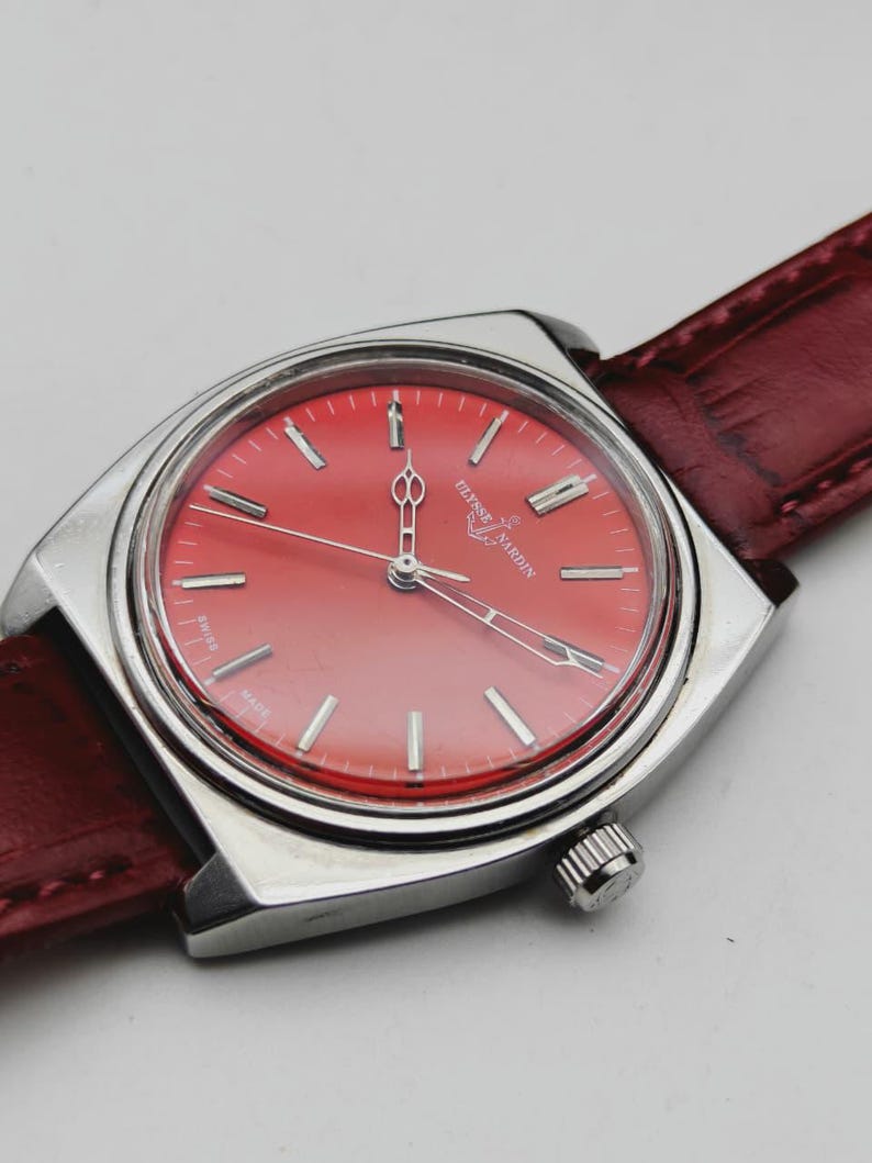 May include: A vintage Ulysse Nardin wristwatch with a vibrant red dial and silver-tone case. The watch features a red leather band and a classic design. The dial displays silver hour markers and hands. The watch is a stylish accessory.