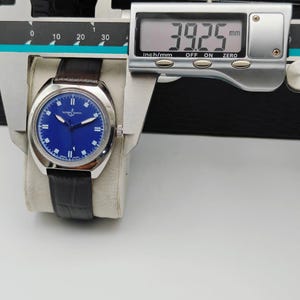 May include: A silver-toned wristwatch with a blue dial and a dark brown leather strap. The watch is measured with a digital caliper, showing a width of 39.25 mm. The watch is resting on a white surface.