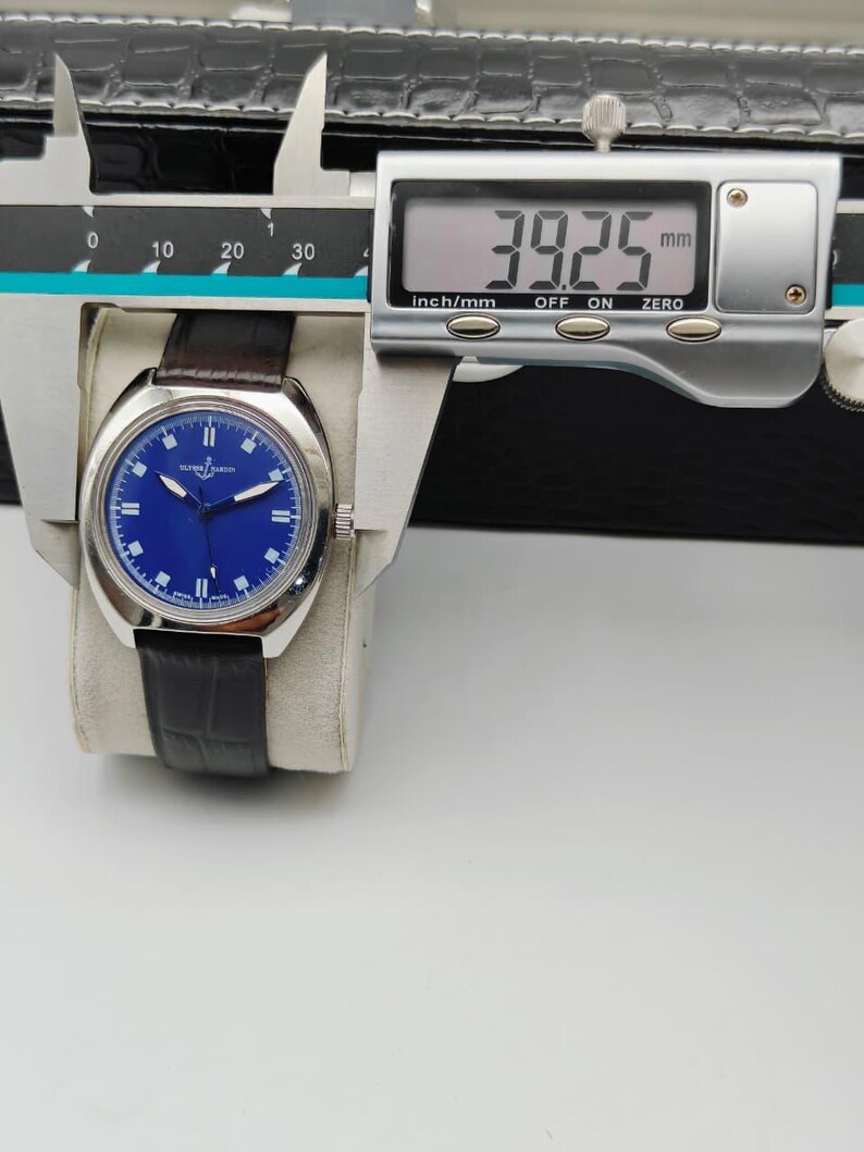 May include: A silver-toned wristwatch with a blue dial and a dark brown leather strap. The watch is measured with a digital caliper, showing a width of 39.25 mm. The watch is resting on a white surface.