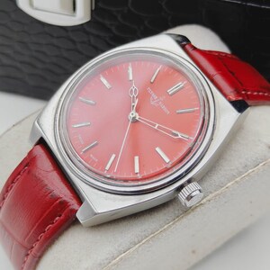 May include: A vintage wristwatch with a vibrant red face and a matching red leather band. The watch has a silver-colored case and features the brand name "Ulysse Nardin" on the face. The watch is displayed on a light-colored surface.