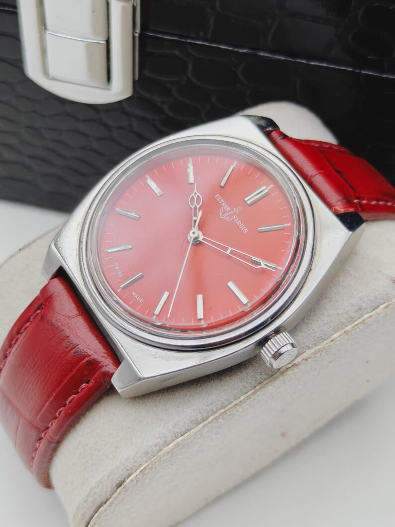 May include: A vintage wristwatch with a vibrant red face and a matching red leather band. The watch has a silver-colored case and features the brand name "Ulysse Nardin" on the face. The watch is displayed on a light-colored surface.