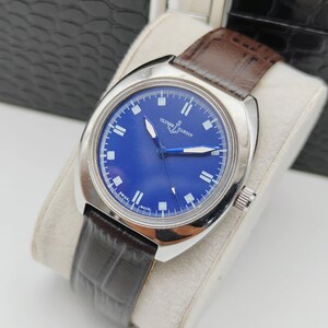 May include: A silver-toned wristwatch with a deep blue face and a brown leather band. The watch face features white markers and hands, with the brand name "Ulysse Nardin" visible. The band is dark brown with a textured pattern.