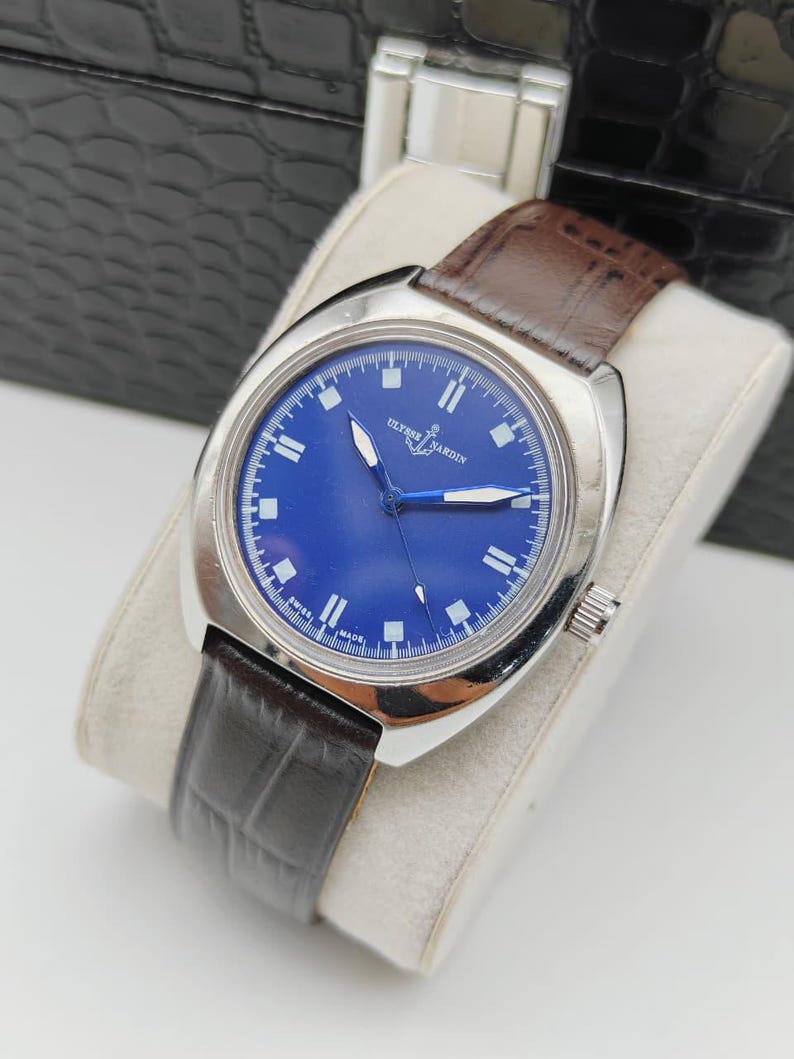 May include: A silver-toned wristwatch with a deep blue face and a brown leather band. The watch face features white markers and hands, with the brand name "Ulysse Nardin" visible. The band is dark brown with a textured pattern.