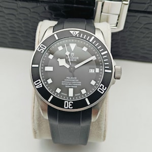 May include: A black and silver Tudor Pelagos watch with a black rubber strap. The watch face is dark gray with white markers and hands. The bezel is black with white numerals. The watch is displayed on a cream-colored cushion.