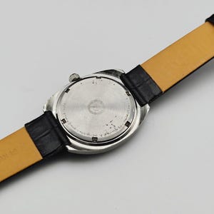 May include: Close-up of the back of a silver-toned wristwatch with a black leather strap. The watch face is circular with a textured design. The strap is black on the outside and tan on the inside.