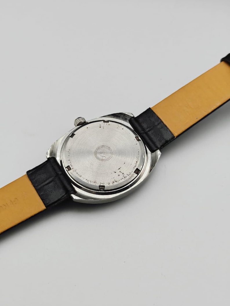 May include: Close-up of the back of a silver-toned wristwatch with a black leather strap. The watch face is circular with a textured design. The strap is black on the outside and tan on the inside.