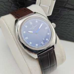 May include: A silver-toned wristwatch with a dark blue face and a brown leather band. The watch has silver hands and markers, and the band is textured. The watch is displayed on a white cushion.