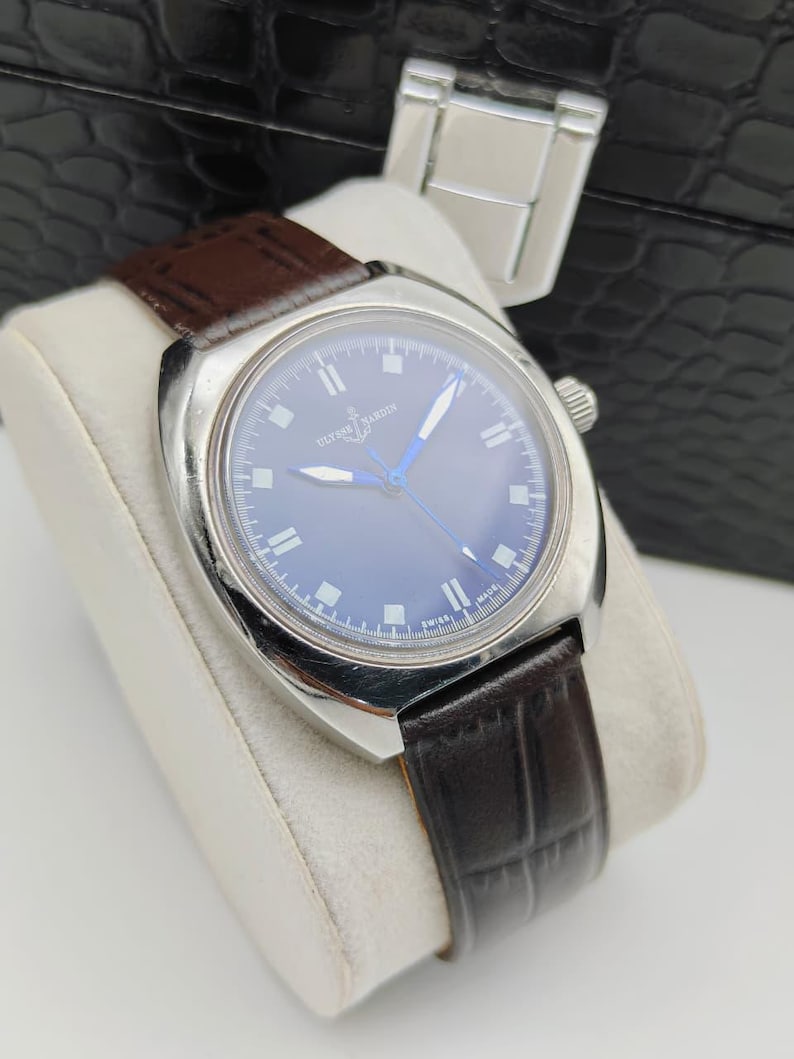 May include: A silver-toned wristwatch with a dark blue face and a brown leather band. The watch has silver hands and markers, and the band is textured. The watch is displayed on a white cushion.