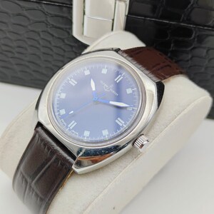 May include: A silver-toned wristwatch with a blue face and brown leather band. The watch has a round face with silver markers and hands. The band is textured and the watch is displayed on a white cushion.