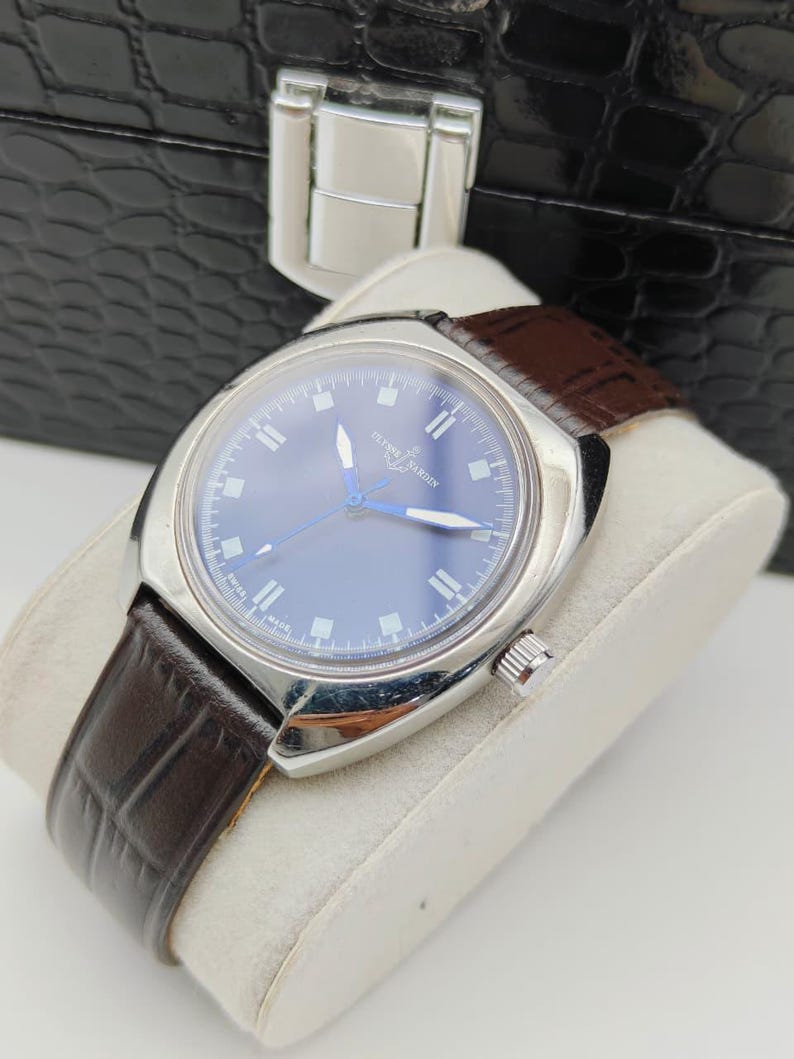 May include: A silver-toned wristwatch with a blue face and brown leather band. The watch has a round face with silver markers and hands. The band is textured and the watch is displayed on a white cushion.