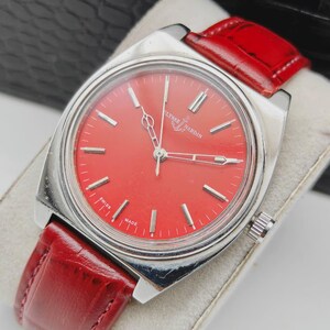May include: A vintage Ulysse Nardin watch with a vibrant red dial and silver-tone case. The watch features a red leather strap and the words "SWISS MADE" are visible on the dial. The watch is displayed on a white surface.