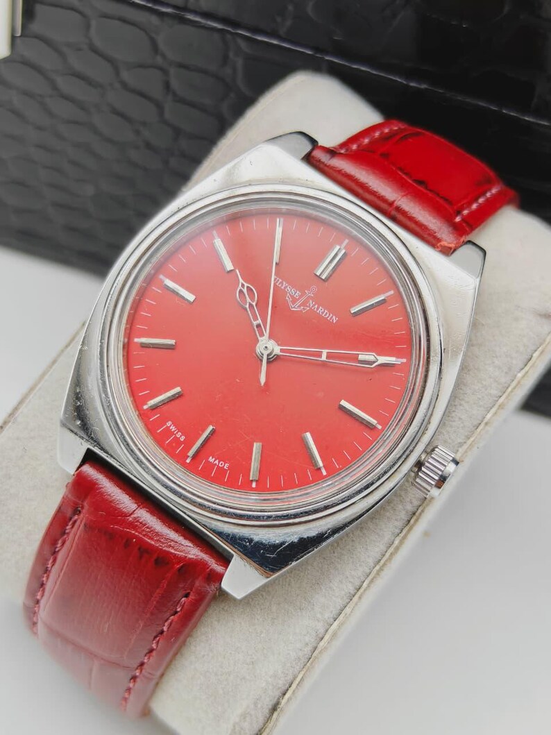 May include: A vintage Ulysse Nardin watch with a vibrant red dial and silver-tone case. The watch features a red leather strap and the words "SWISS MADE" are visible on the dial. The watch is displayed on a white surface.