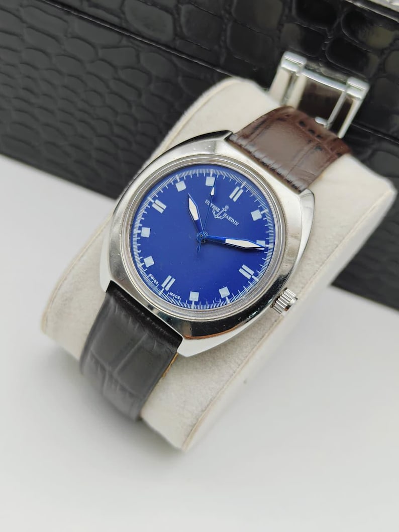 May include: A vintage Ulysse Nardin watch with a striking blue face and silver casing. The watch features white hour markers and hands, complemented by a brown leather strap. The watch is displayed on a white cushion.