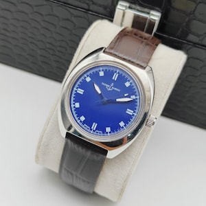 May include: A silver-toned wristwatch with a blue face and a brown leather band. The watch face features white hour markers and hands, with the brand name "ULYSSE NARDIN" printed on the face. The watch is displayed on a white cushion.