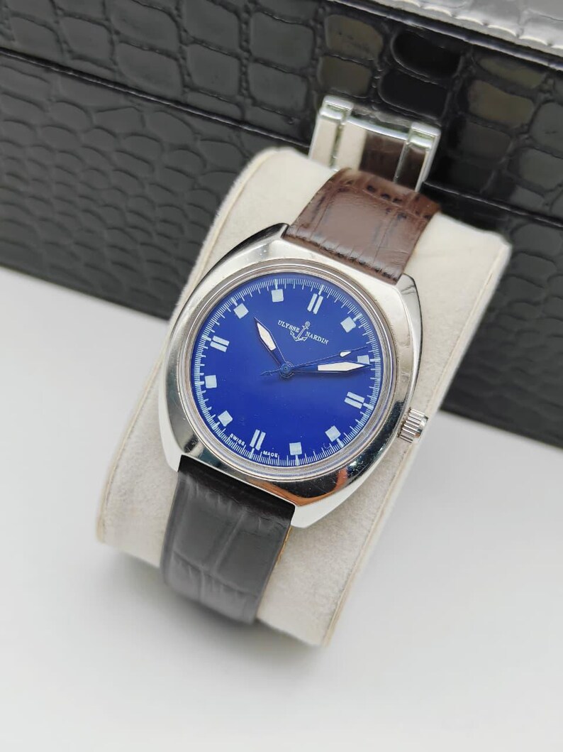 May include: A silver-toned wristwatch with a blue face and a brown leather band. The watch face features white hour markers and hands, with the brand name "ULYSSE NARDIN" printed on the face. The watch is displayed on a white cushion.
