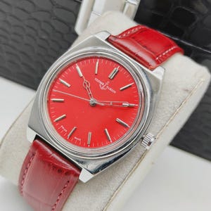 May include: A vintage wristwatch with a vibrant red face and silver-toned case. The watch features a red leather band and the brand name "Ulysse Nardin" is visible on the face. The watch is displayed on a white surface.