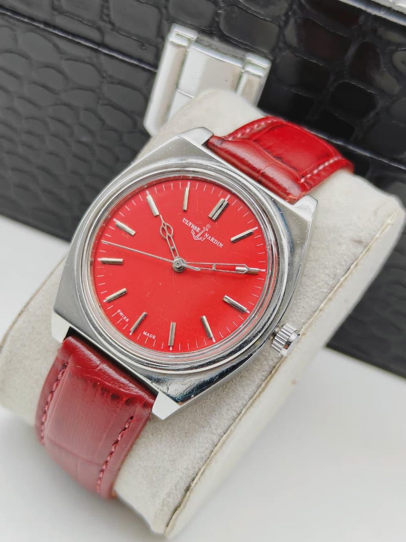 May include: A vintage wristwatch with a vibrant red face and silver-toned case. The watch features a red leather band and the brand name "Ulysse Nardin" is visible on the face. The watch is displayed on a white surface.