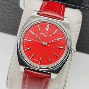 May include: A vintage Ulysse Nardin wristwatch with a vibrant red dial and silver-tone case. The watch features a red leather strap and is marked "Swiss Made". The watch is displayed on a cream-colored cushion.