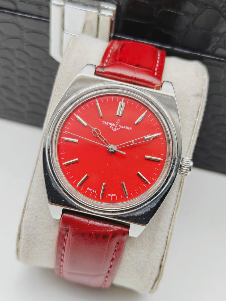 May include: A vintage Ulysse Nardin wristwatch with a vibrant red dial and silver-tone case. The watch features a red leather strap and is marked "Swiss Made". The watch is displayed on a cream-colored cushion.