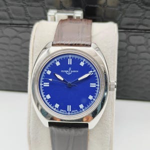 May include: A silver-toned wristwatch with a deep blue face and a brown leather strap. The watch face features white hour markers and hands, with the brand name "ULYSSE NARDIN" visible. The watch is displayed on a white cushion.