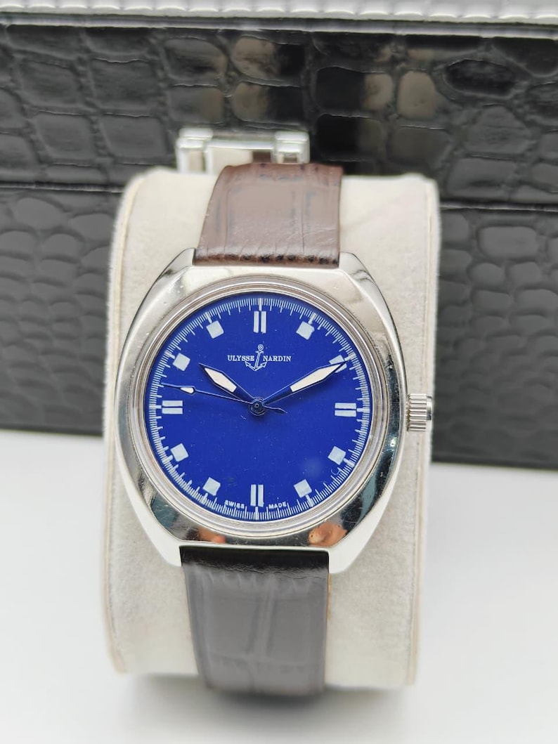 May include: A silver-toned wristwatch with a deep blue face and a brown leather strap. The watch face features white hour markers and hands, with the brand name "ULYSSE NARDIN" visible. The watch is displayed on a white cushion.