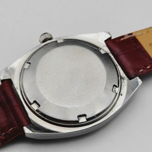 May include: Close-up of the back of a silver-toned wristwatch with a burgundy leather strap. The watch case is rectangular with a circular back. The strap shows signs of wear.