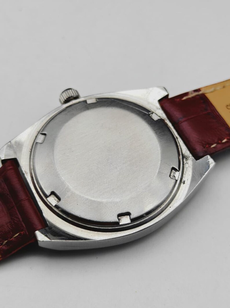 May include: Close-up of the back of a silver-toned wristwatch with a burgundy leather strap. The watch case is rectangular with a circular back. The strap shows signs of wear.