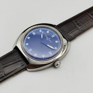 May include: A silver-toned Ulysse Nardin wristwatch with a blue face and a brown leather band. The watch has a round face with white hour markers and blue hands. The band is textured and has a buckle closure.