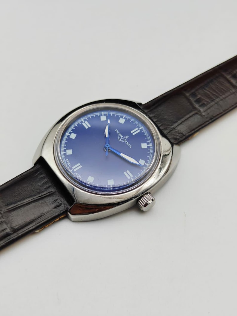 May include: A silver-toned Ulysse Nardin wristwatch with a blue face and a brown leather band. The watch has a round face with white hour markers and blue hands. The band is textured and has a buckle closure.