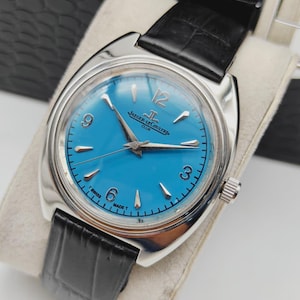 Puede incluir: Un reloj Jaeger-LeCoultre Club vintage con una esfera azul vibrante y agujas e indicadores plateados. El reloj presenta una caja de acero inoxidable y una correa de cuero negro. El reloj se muestra sobre un cojín color crema.