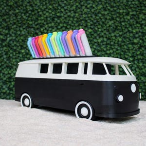 May include: A black and white van-shaped desk organizer with a row of colorful pens on top. The van has white wheels and headlights. The background is a green hedge.