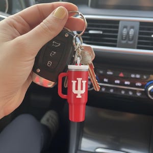 Op de afbeelding: Rode miniatuur tumbler sleutelhanger met een witte deksel en handvat. De sleutelhanger heeft het Indiana University trident logo in het wit. De sleutelhanger is bevestigd aan een sleutelring met autosleutels en huissleutels.