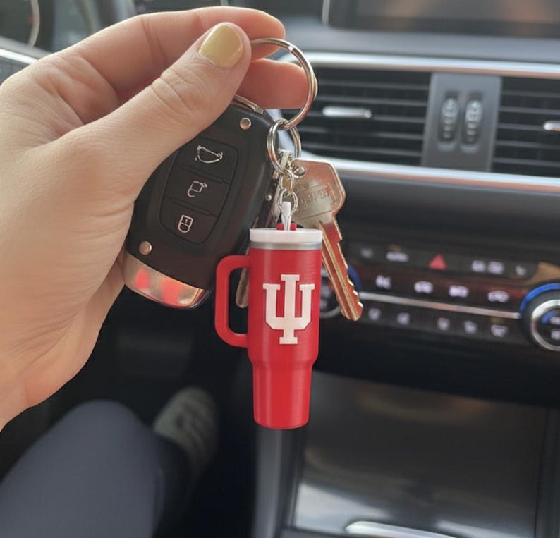 Op de afbeelding: Rode miniatuur tumbler sleutelhanger met een witte deksel en handvat. De sleutelhanger heeft het Indiana University trident logo in het wit. De sleutelhanger is bevestigd aan een sleutelring met autosleutels en huissleutels.