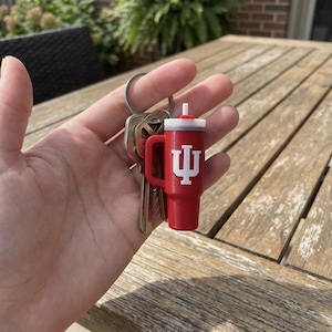 Op de afbeelding: Een rode sleutelhanger in de vorm van een beker met een wit deksel en handvat, met een wit logo. De sleutelhanger is bevestigd aan een zilveren sleutelring met meerdere sleutels.