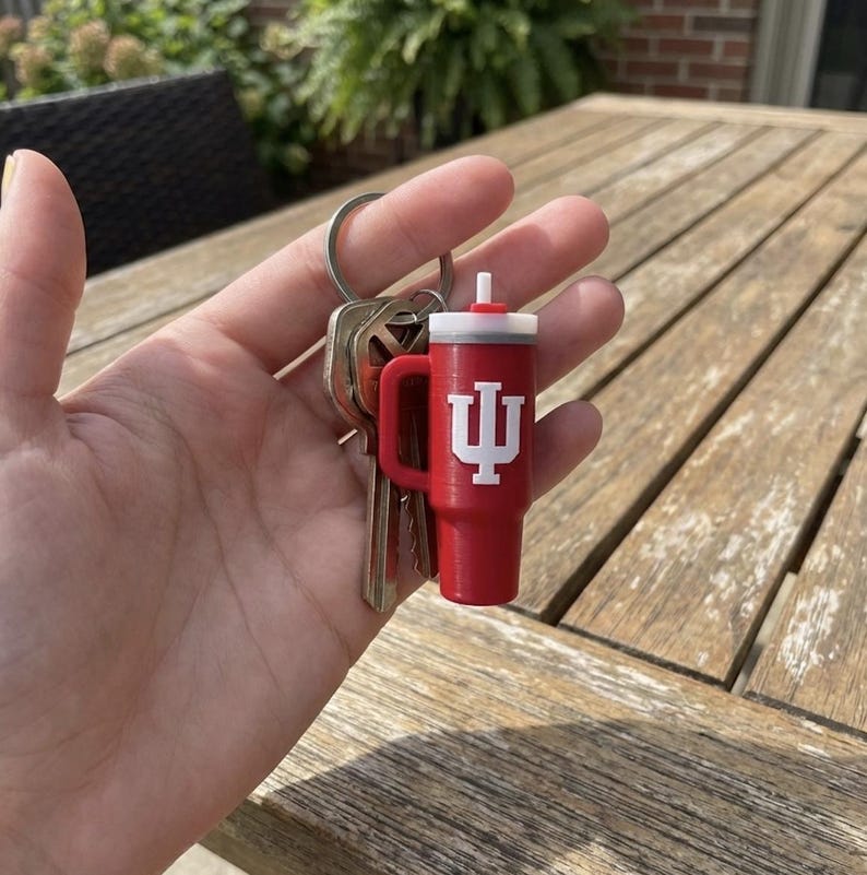 Op de afbeelding: Een rode sleutelhanger in de vorm van een beker met een wit deksel en handvat, met een wit logo. De sleutelhanger is bevestigd aan een zilveren sleutelring met meerdere sleutels.